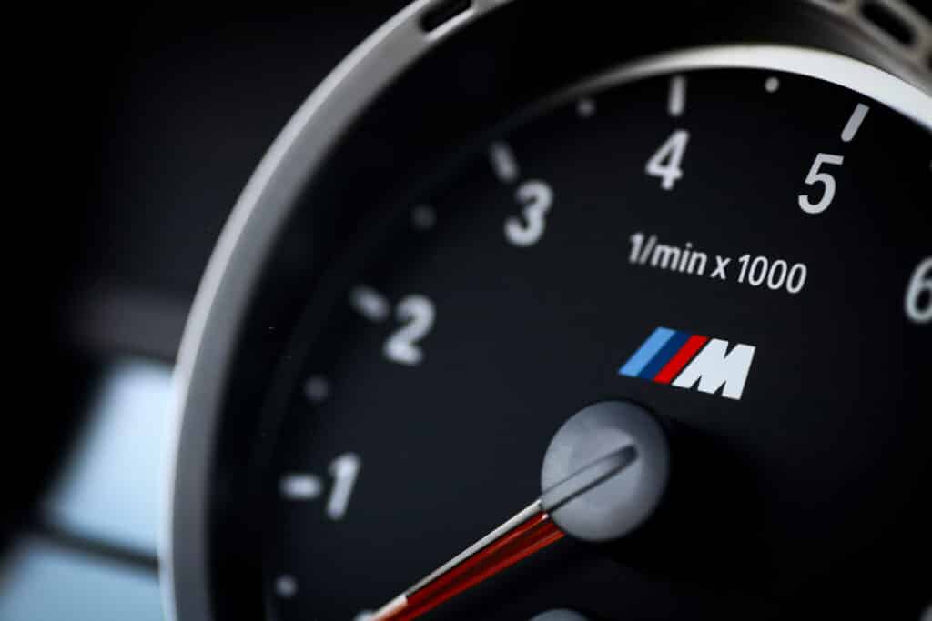 Close-up shot of a BMW M series tachometer (rev counter) gauge, showing the prominent BMW 'M' logo with the red, violet, and blue stripes on a black background.