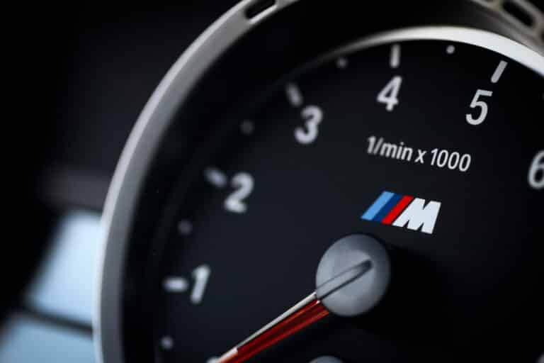 Close-up shot of a BMW M series tachometer (rev counter) gauge, showing the prominent BMW 'M' logo with the red, violet, and blue stripes on a black background.