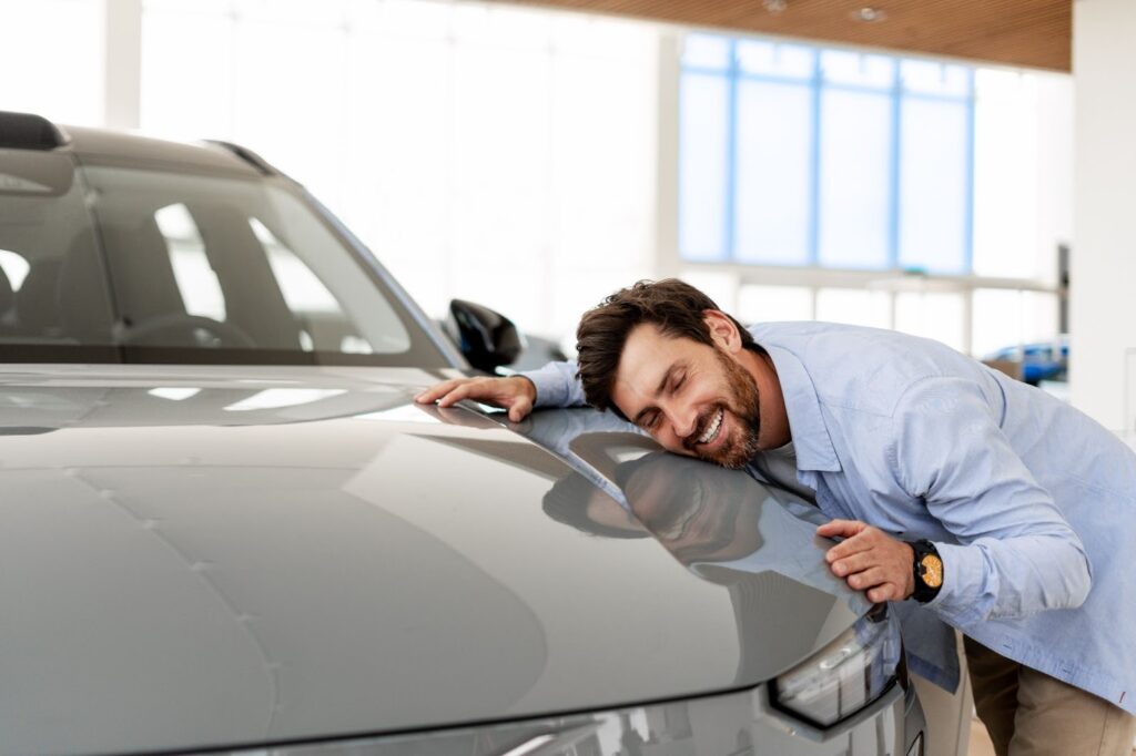 Happy South African man with a beard embracing the bonnet of a new grey SUV in a showroom, smiling with his eyes closed.